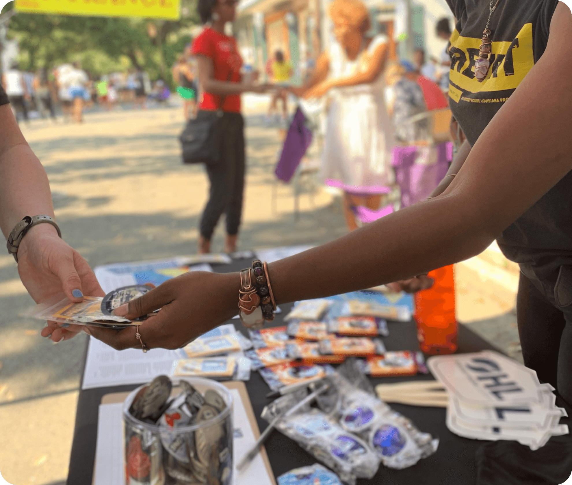 Person receiving items from another at an outdoor event with a table displaying various items.