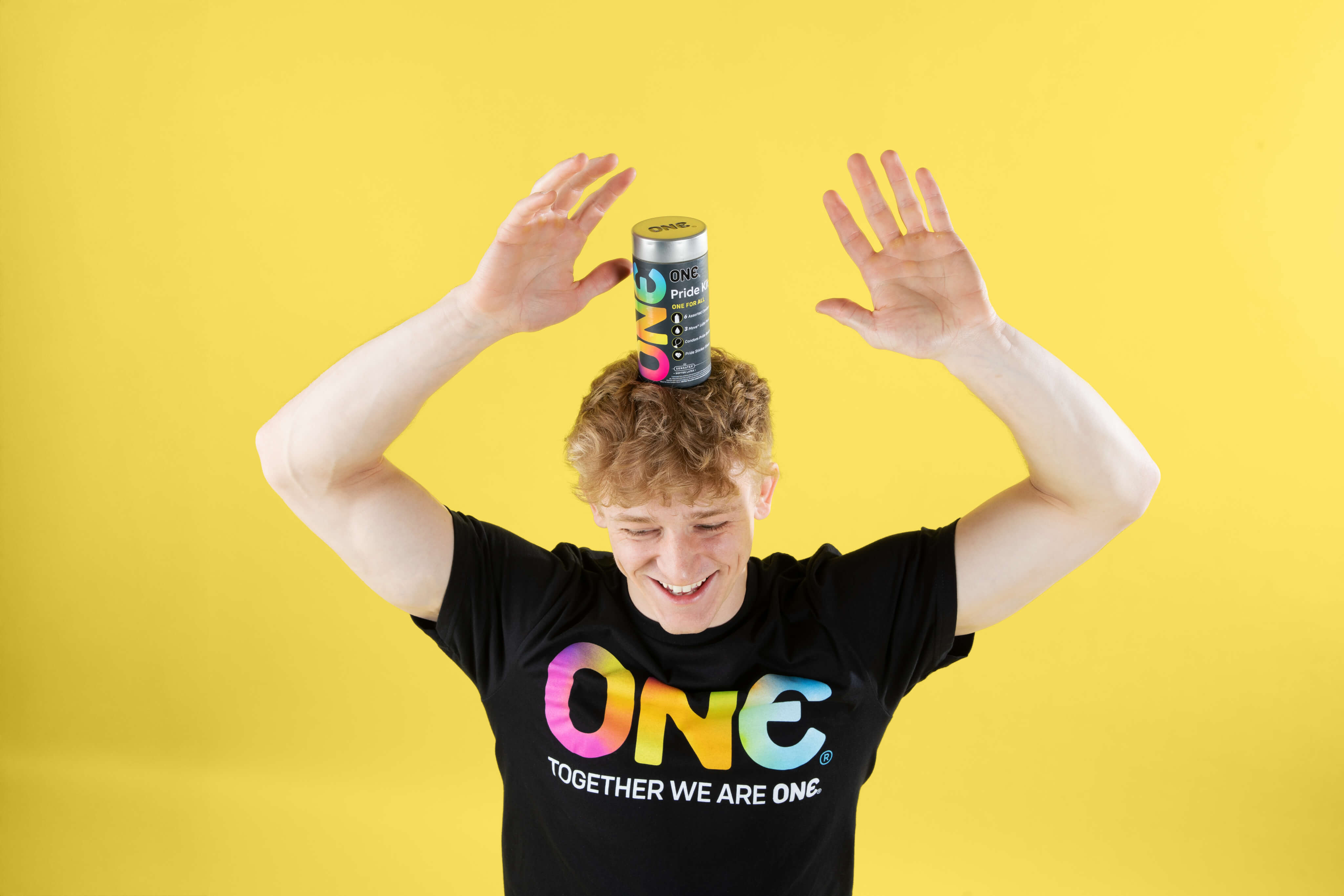 Person wearing a black t-shirt with 'ONE' printed on it, holding a colorful can above their head against a yellow background.