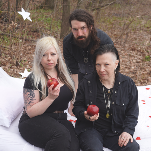 Image of Astral Witch - a new ONE On Stage band. Three people in black in the woods sitting on a white blanket. Two people hold apples.