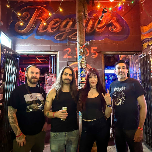 Image of Black Road - a ONE on Stage band. Four members standing in venue. Wall in background says 