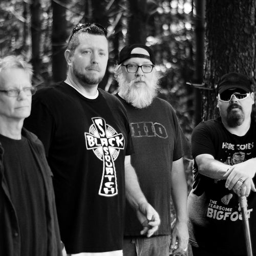 Four men standing in a forest, wearing black t-shirts with visible text and graphics.