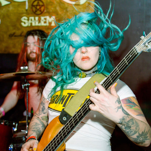 Person with blue hair playing a bass guitar in a band setting.