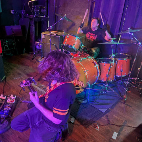 Two musicians playing guitar and drums on a stage with purple lighting.