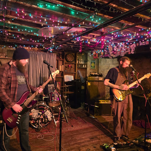 Image of two people playing guitars infront of microphones. Colored lights hang on the ceiling above them. 