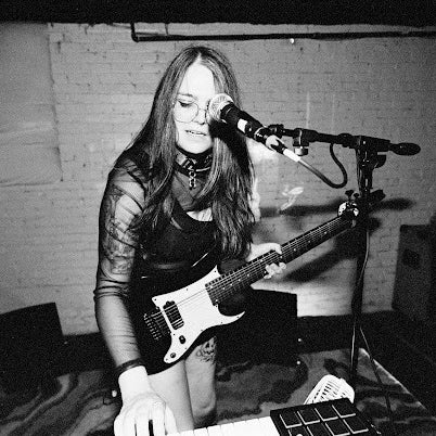 Person playing guitar and singing into a microphone in a room with brick walls.